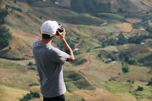 A person with a camera takes a picture of a lush green hillside with tattoo visible, wearing a gray shirt and white cap.