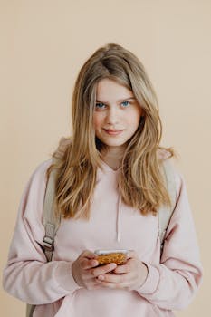 Blonde teenage girl holding mobile phone, wearing a pink hoodie against a plain background.