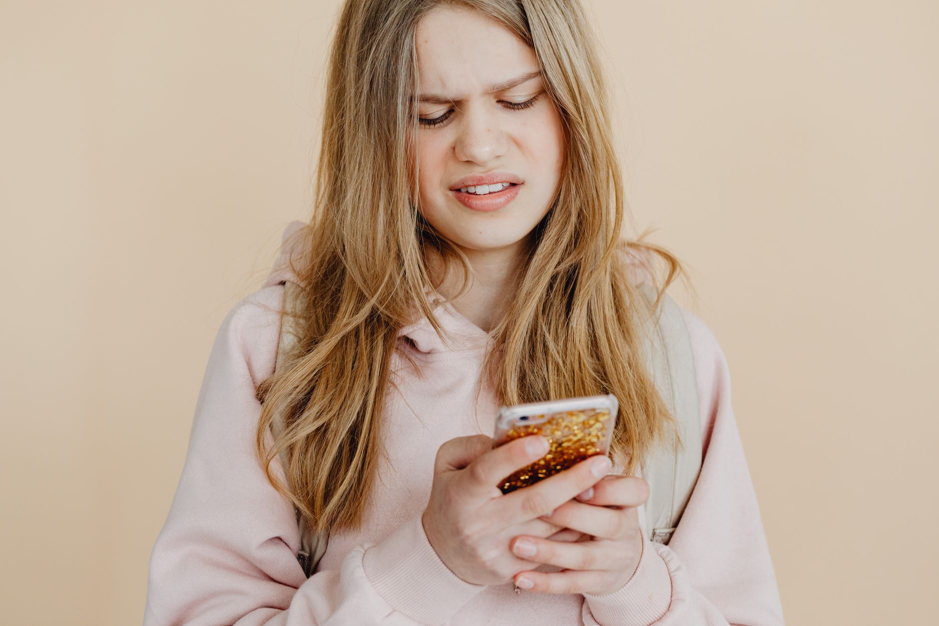 Teen girl with blonde hair in pink hoodie using smartphone, expressing curiosity indoors.