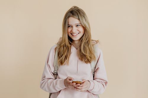 Free A cheerful teenager with blonde hair wearing a pink sweater and using a smartphone indoors. Stock Photo