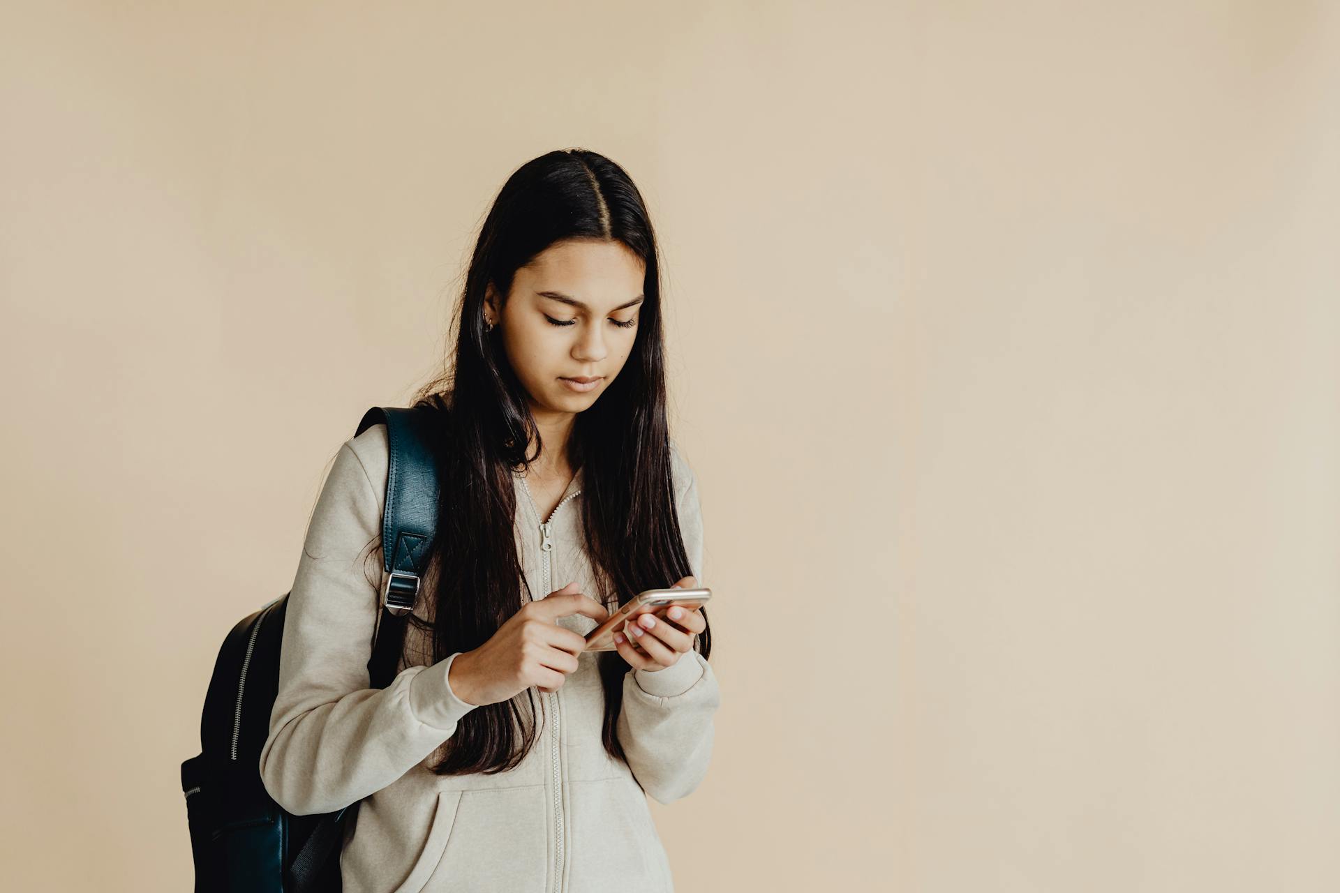 Teenage girl with backpack using a smartphone in a minimalist indoor setting, suited for educational themes.