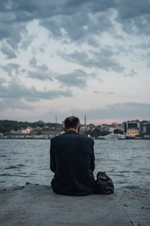Back view of unrecognizable male in suit sitting on stone pier near rippling water and admiring view of city in evening time