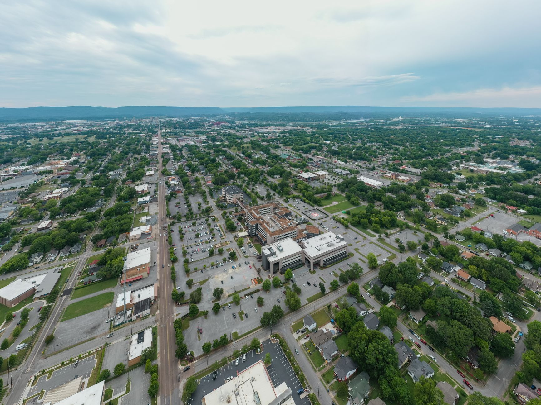 Aerial view of a sprawling urban area with green spaces and distant mountains under a cloudy sky.