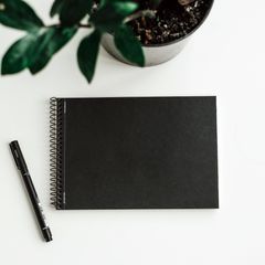 A small black spiral-bound sketchbook with a fineliner beside a plant