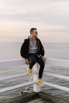 Man in casual attire sitting on Sopot Pier railing with ocean backdrop during a serene day.