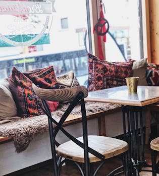 A charming cafe interior with vintage pillows, wooden chairs, and a window view.
