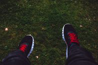 View of feet in black sneakers with red laces hovering above green grass.