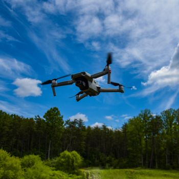 Capture of a drone flying above a lush green forest under a bright blue sky.