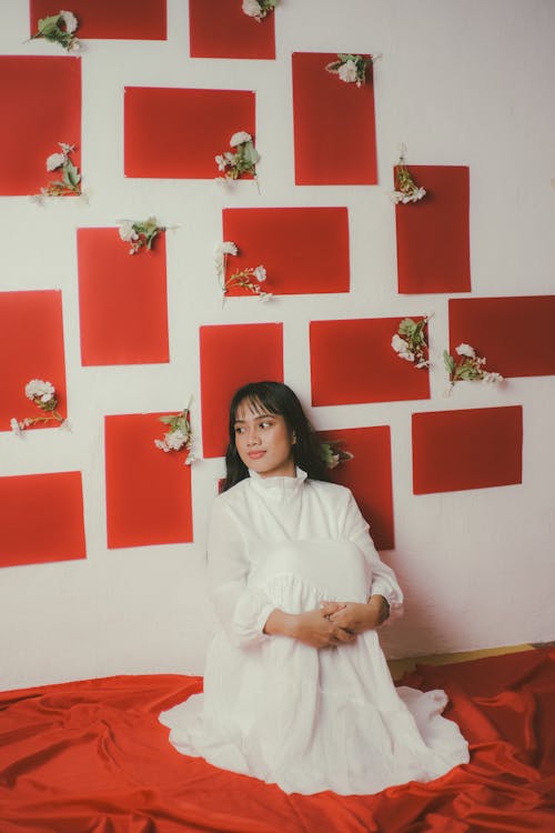 Woman in White Dress Sitting by the Wall on Red Sheets 