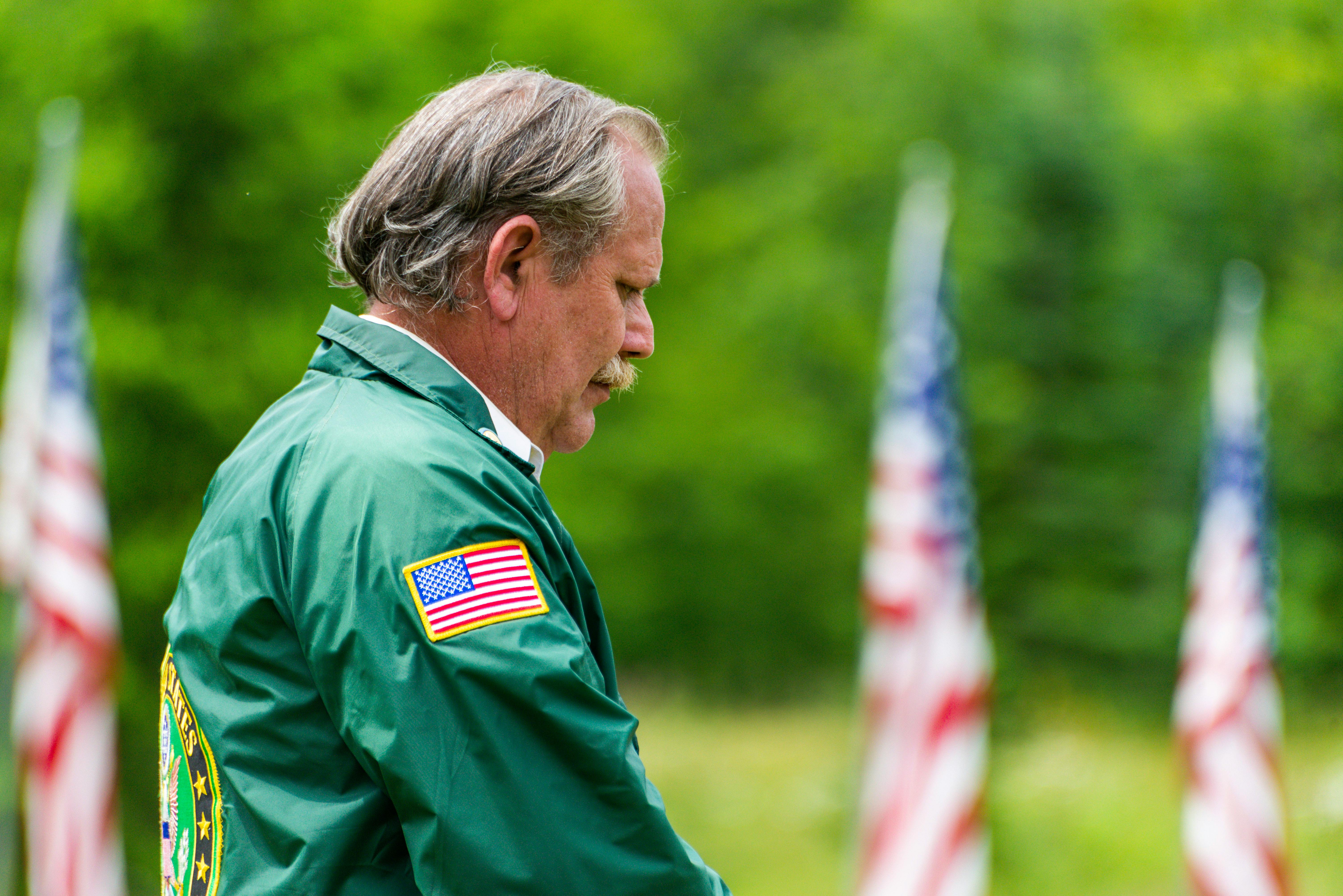 Senior adult standing outdoors, wearing a green jacket with USA flag, surrounded by American flags in a park setting.