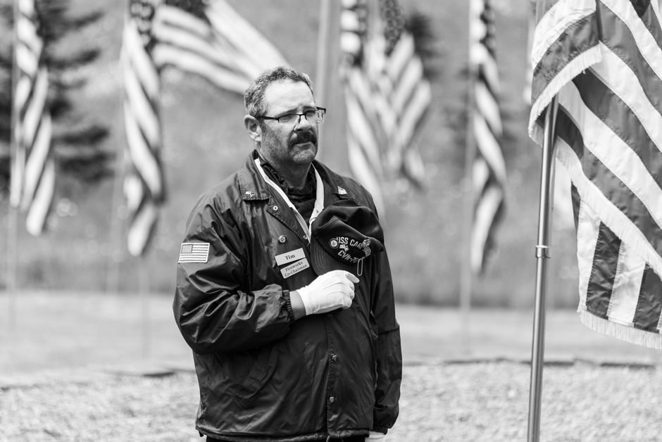 A veteran holding a hat stands solemnly among flags at a military memorial in black and white. A veteran holding a hat stands solemnly among flags at a military memorial in black and white.
