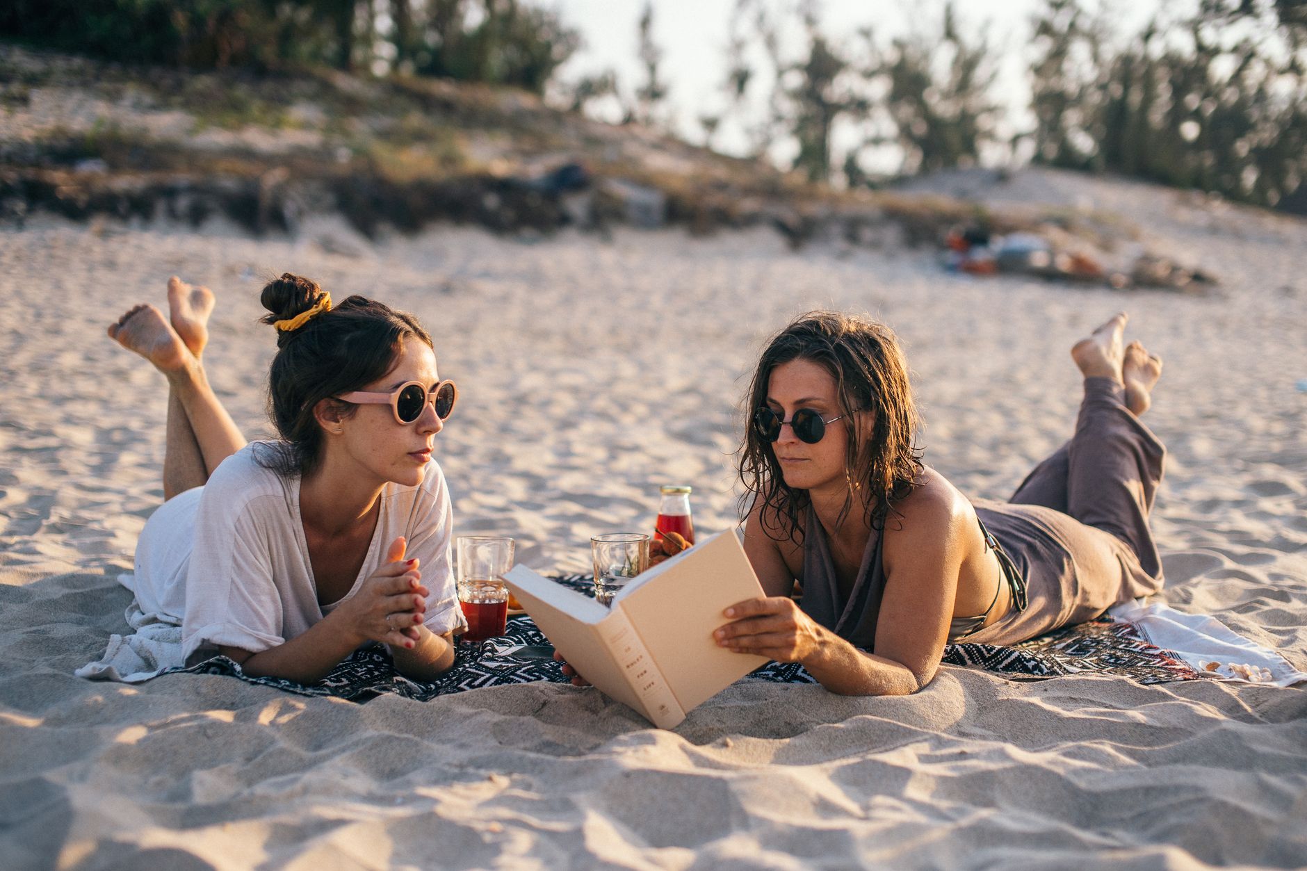 Two women relaxing on a Vietnamese beach, reading and enjoying drinks during a sunny day.
