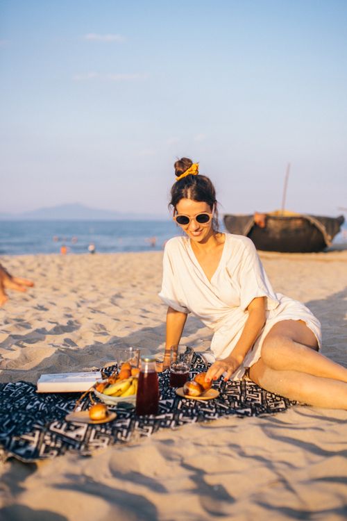 Free Woman relaxing on the beach with a picnic blanket during sunset, wearing sunglasses and smiling. Stock Photo
