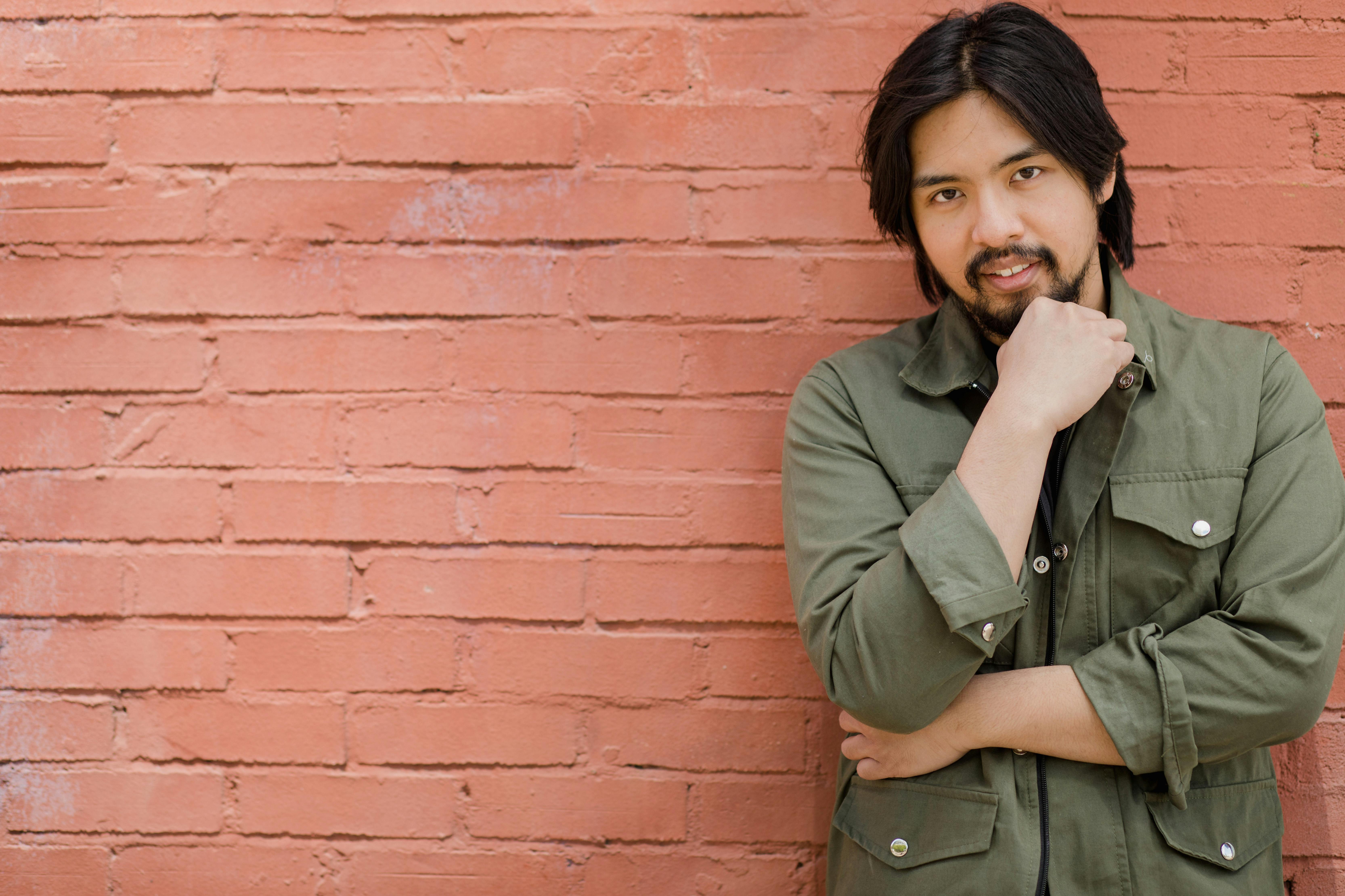 Portrait of a smiling Asian man in green jacket posing against a brick wall.