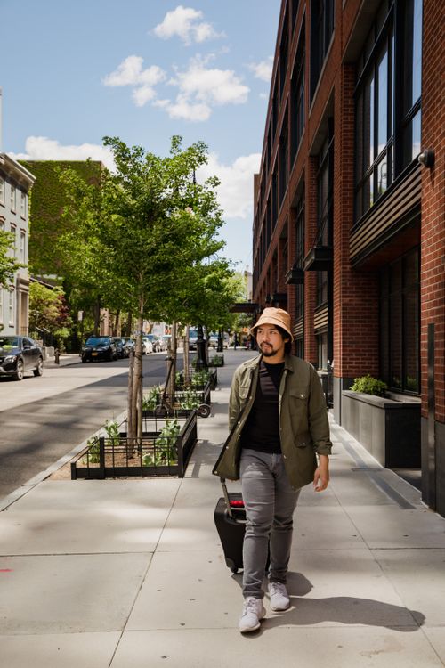Free Bearded man walking along a modern city street pulling luggage under a clear sky. Stock Photo
