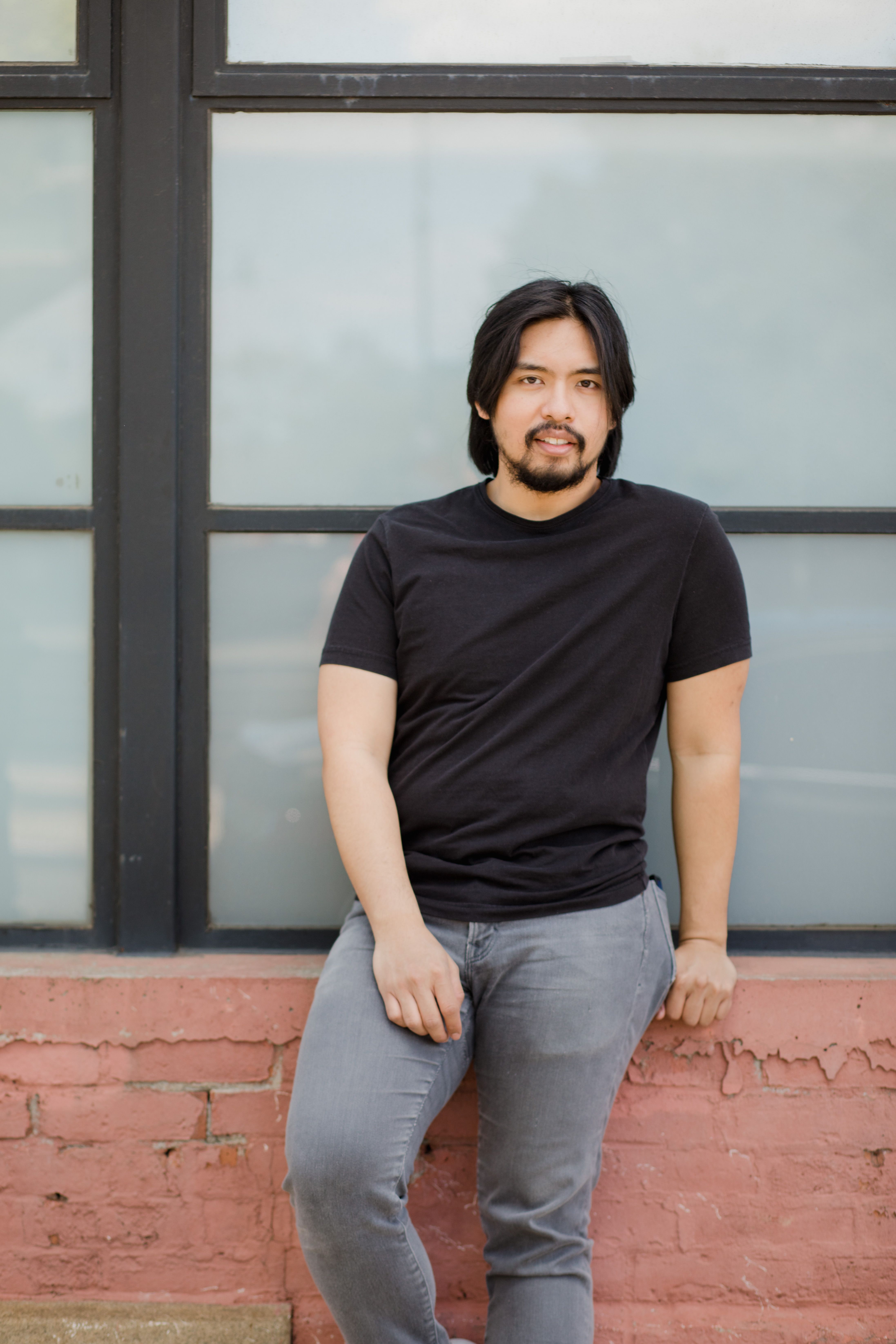 Relaxed man in black t-shirt sitting by a large window with a brick wall background.