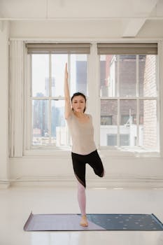 A woman practices yoga indoors in a bright room with cityscape windows, embodying calm and focus.