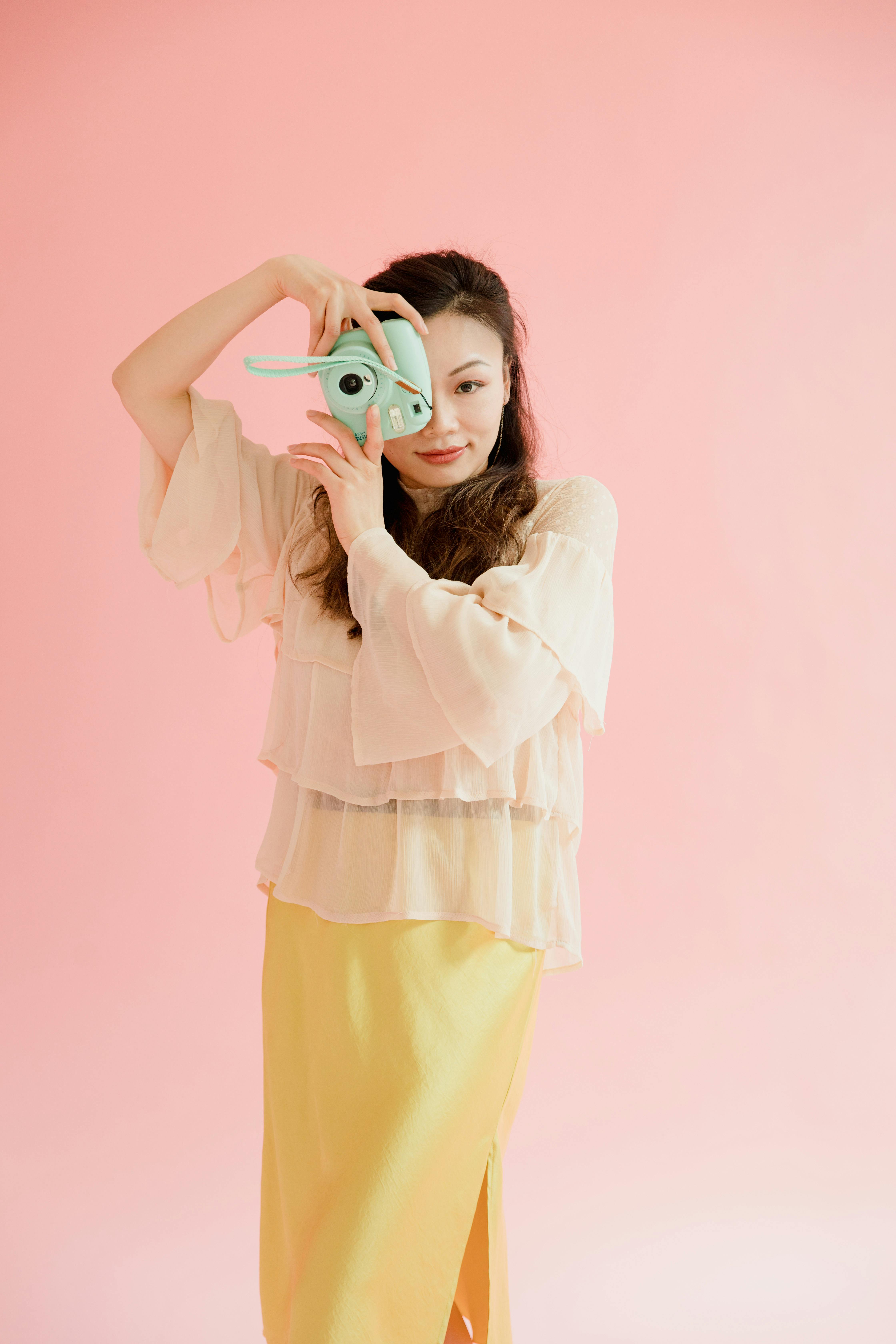 Stylish portrait of an Asian woman posing with a polaroid camera against a pink background.