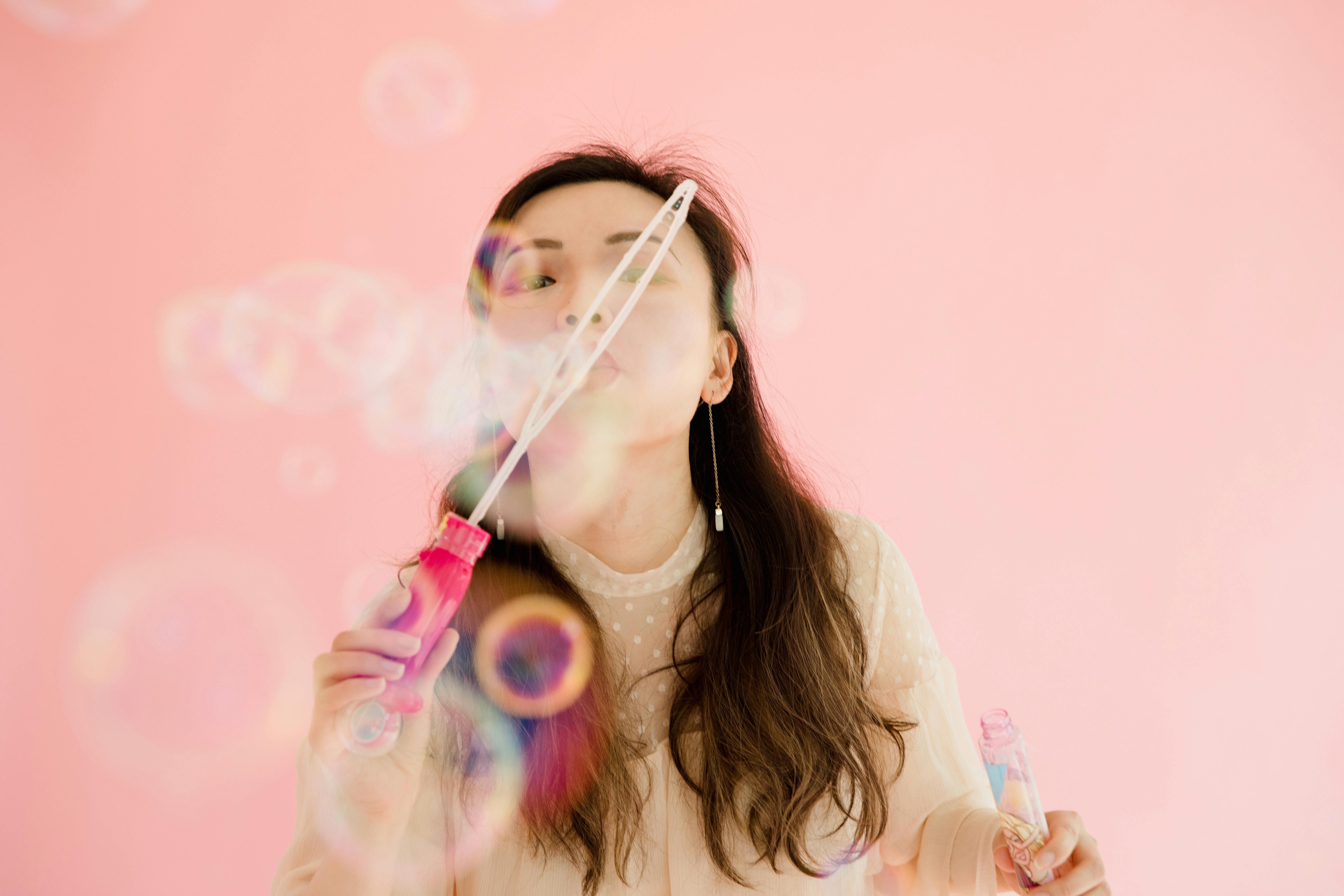 A beautiful Asian woman playfully blowing bubbles in a pink background studio.