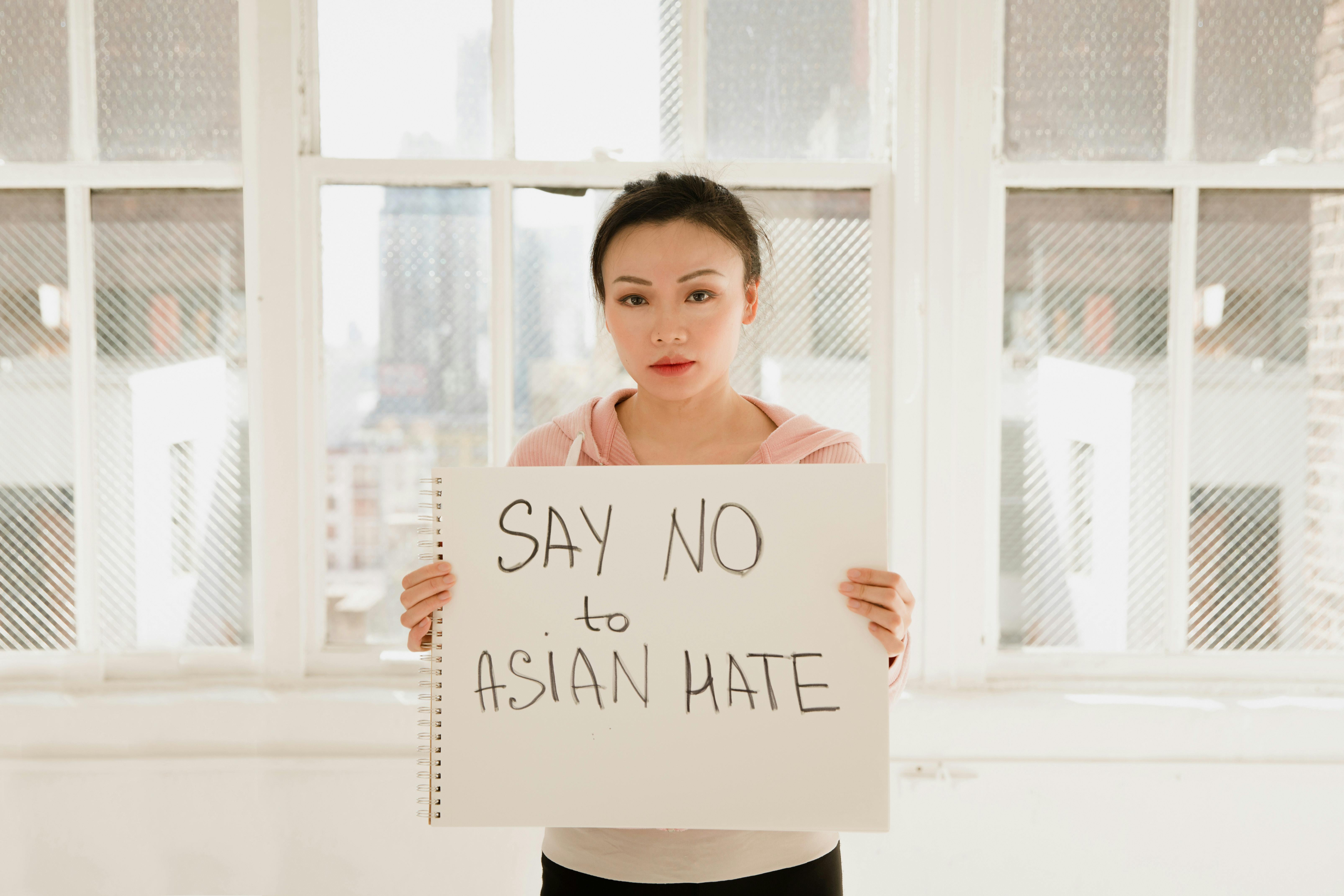An Asian woman holds a sign reading 'Say No to Asian Hate' in a well-lit room.