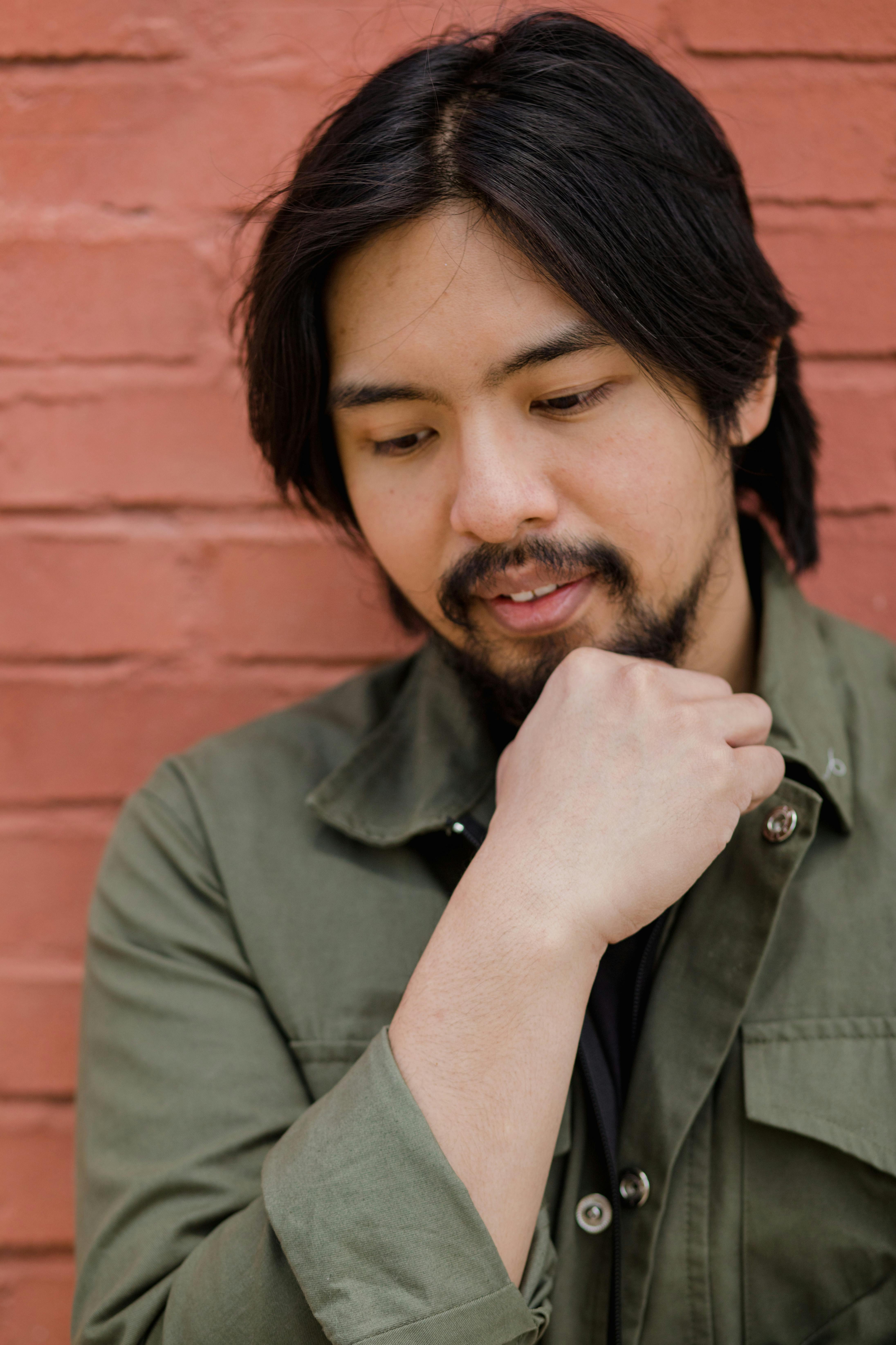 A bearded Asian man in casual green jacket posing thoughtfully against a brick wall.