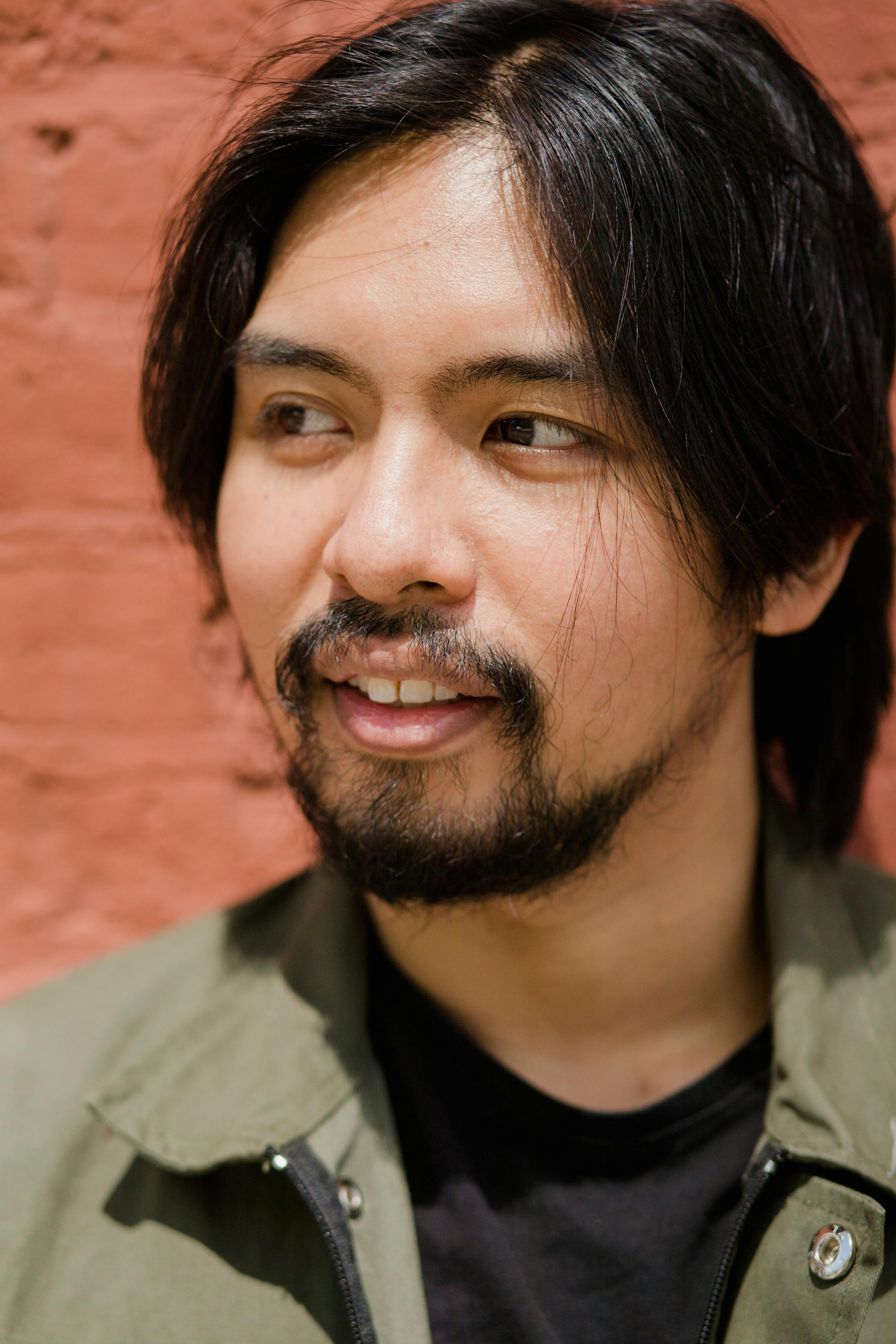 Close-up portrait of a smiling man with facial hair against a brick wall.