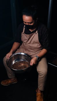 A barista wearing an apron examines coffee beans in a large bowl, seated indoors.