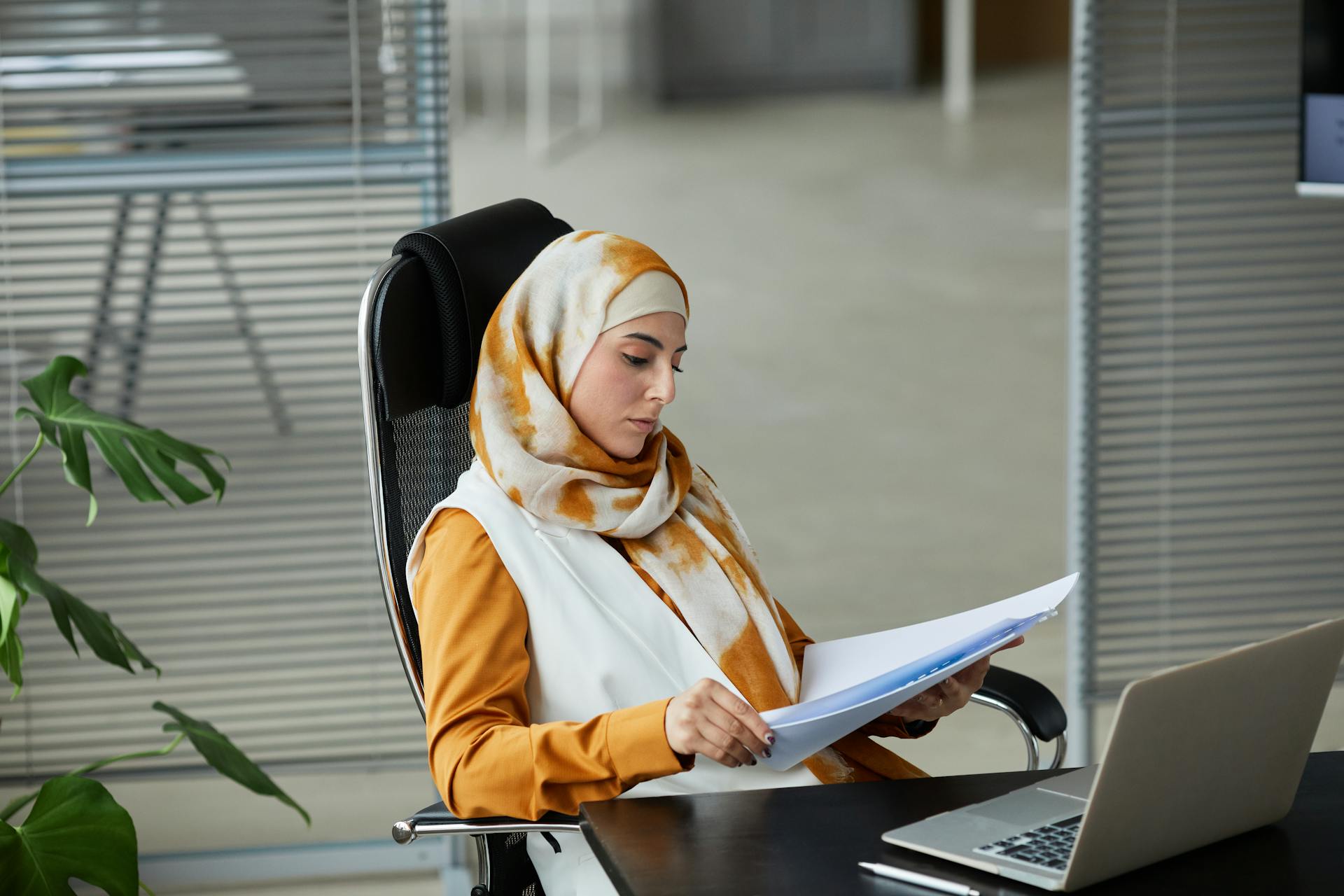 Middle Eastern businesswoman wearing a hijab reading documents at her office desk with a laptop.