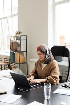A Muslim woman wearing a hijab is focused on her laptop in a modern office setting.