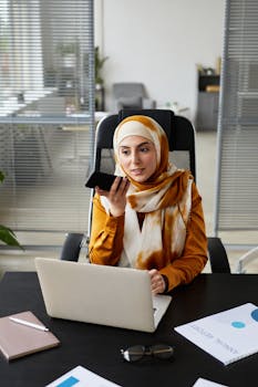 Muslim businesswoman in hijab multitasks with smartphone and laptop at office desk.