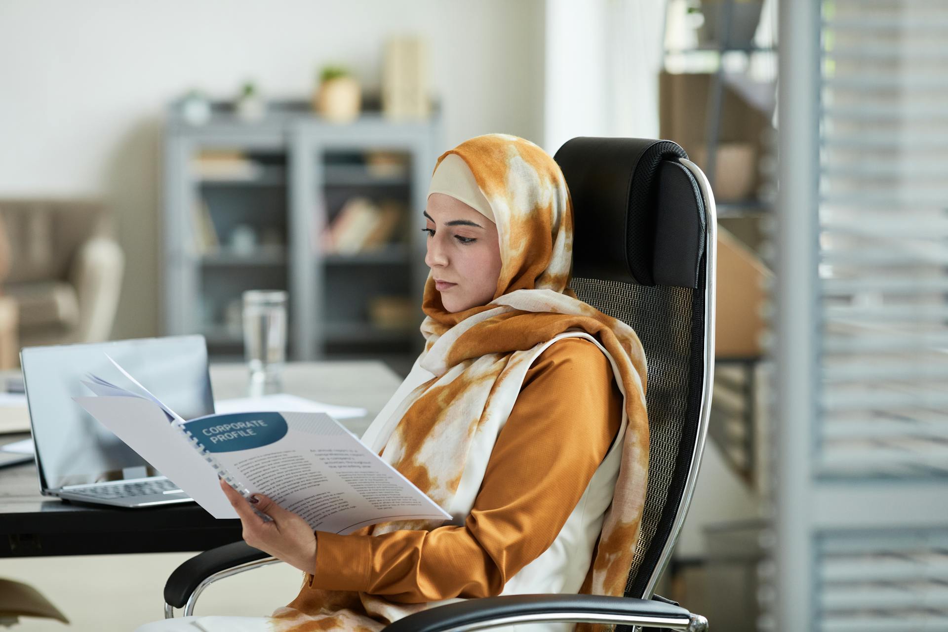 Middle Eastern woman in hijab reading documents at office desk, reflecting focus and professionalism.