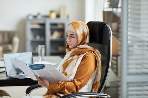 Free Middle Eastern woman in hijab reading documents at office desk, reflecting focus and professionalism. Stock Photo