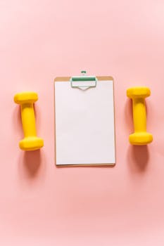Flat lay of yellow dumbbells and a clipboard on a pastel pink background.