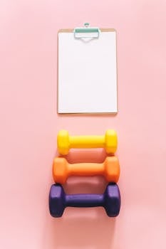 Vibrant dumbbells and clipboard on pink surface, ideal for fitness and exercise themes.