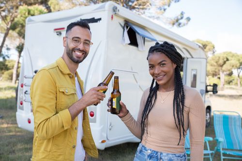 Free Smiling couple holds beer bottles in front of an RV, enjoying a sunny day outdoors. Stock Photo