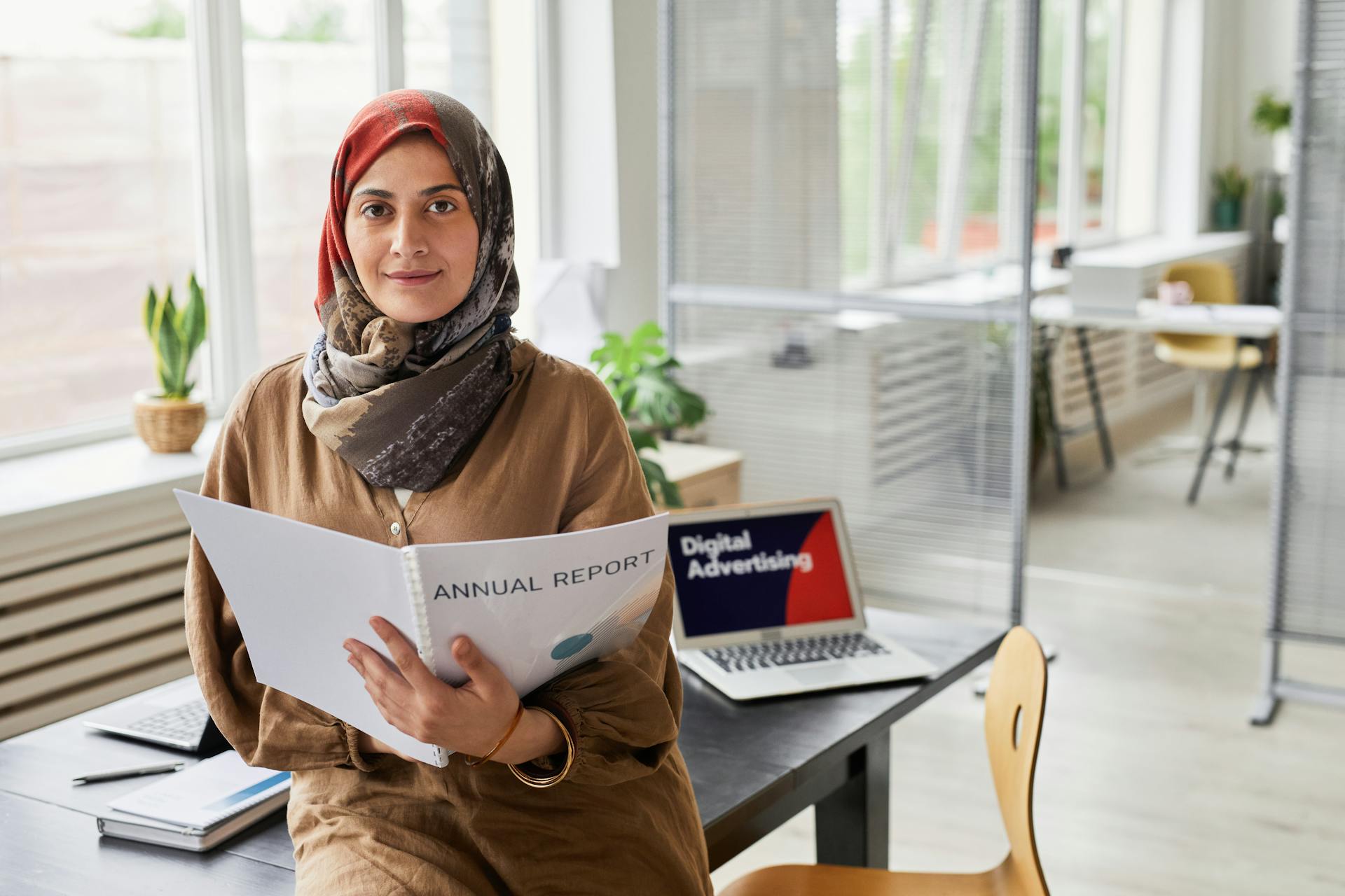 A woman in a hijab holds an annual report in a contemporary office setting.