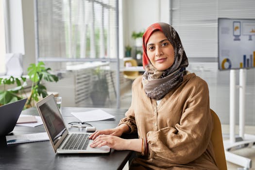 Confident woman in hijab working at office desk with laptop and documents, showcasing productivity.