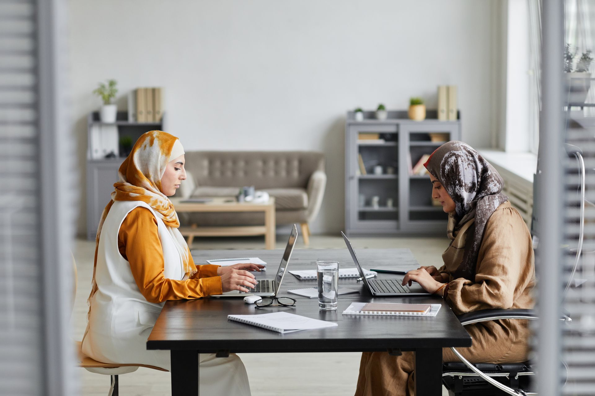 Two women wearing hijabs working on laptops at a modern desk in an office setting.