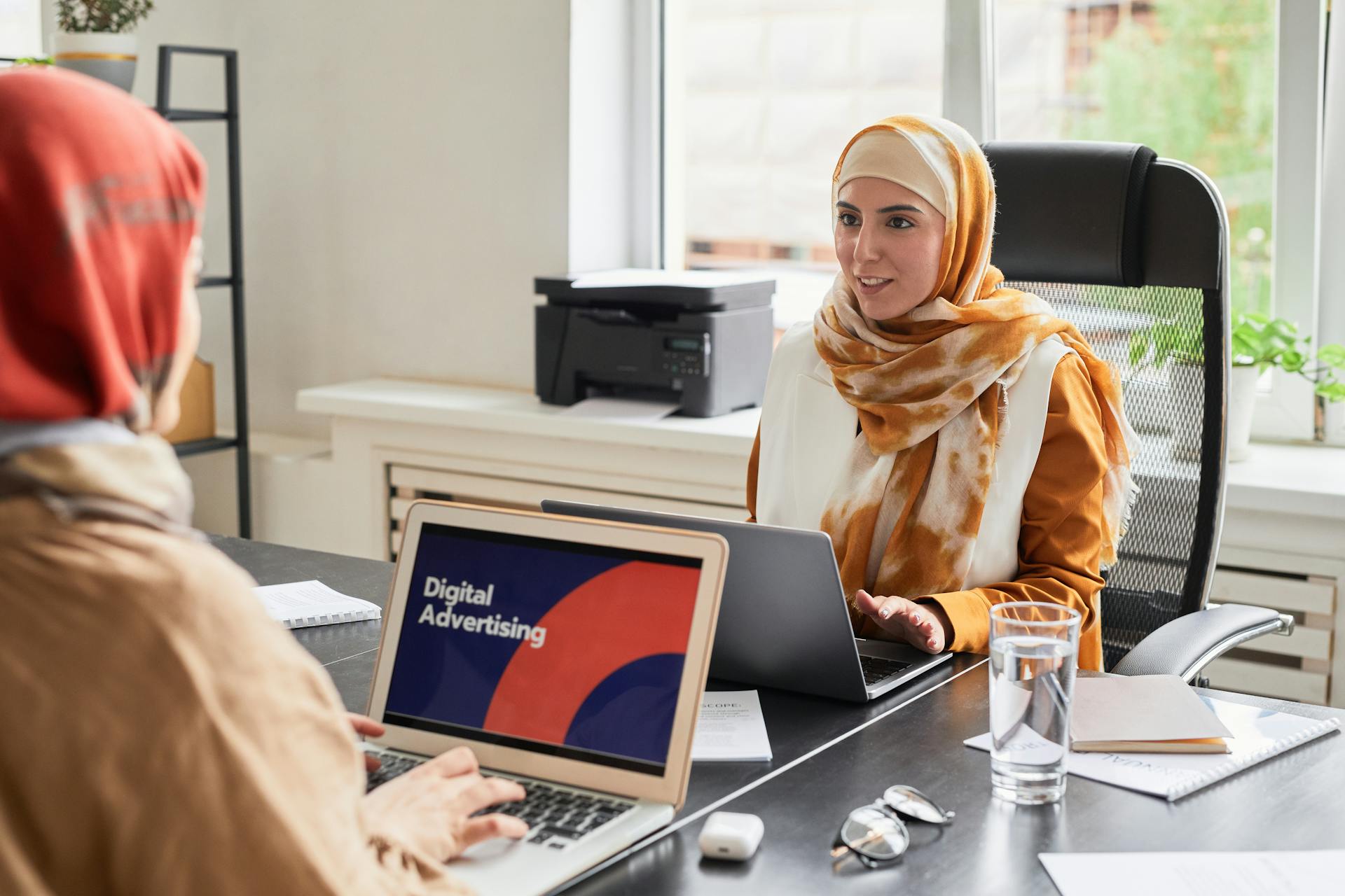 Two women in hijabs working on laptops in an office discussing digital advertising.