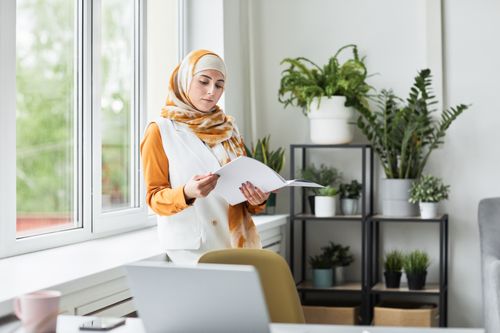 Free A young Muslim woman in hijab reading a document in a modern office with greenery. Stock Photo