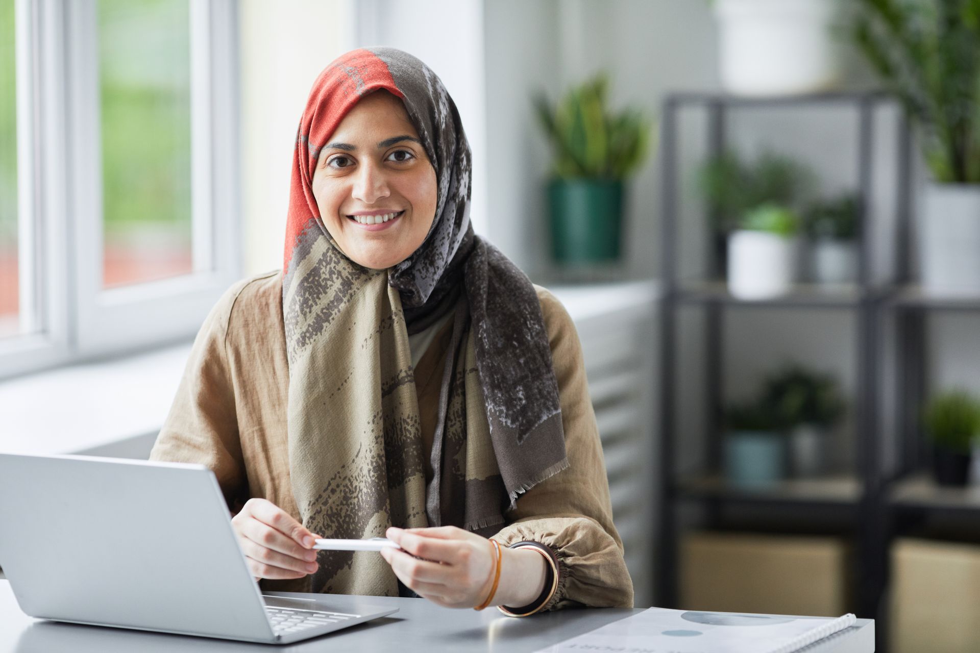 Smiling Muslim woman in hijab working on laptop in a modern office setting.