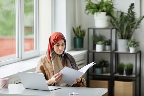 Free Muslim woman in hijab reviewing paperwork at desk with laptop and plants. Stock Photo