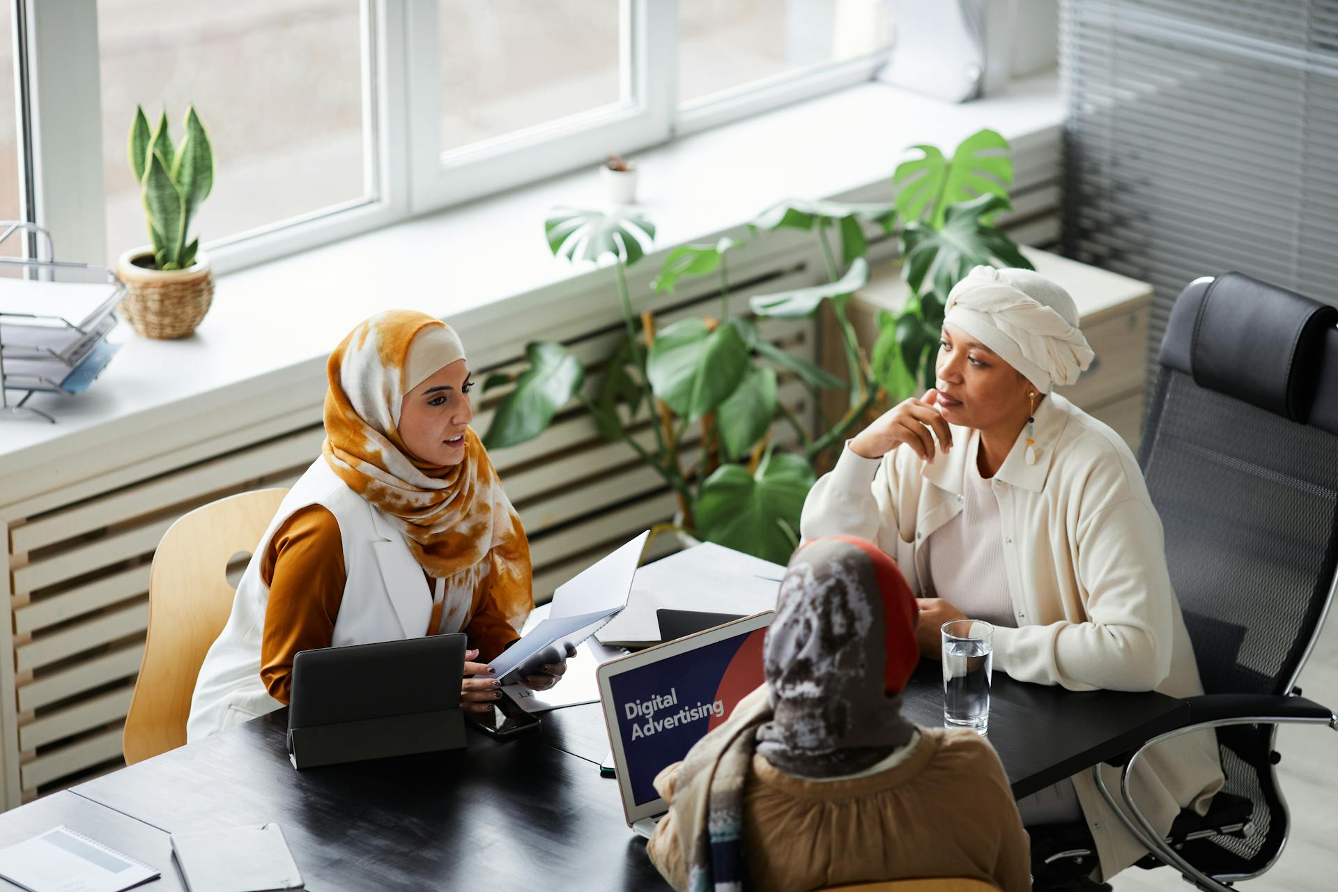 Three diverse women in hijabs discussing digital advertising strategy in a bright modern office.