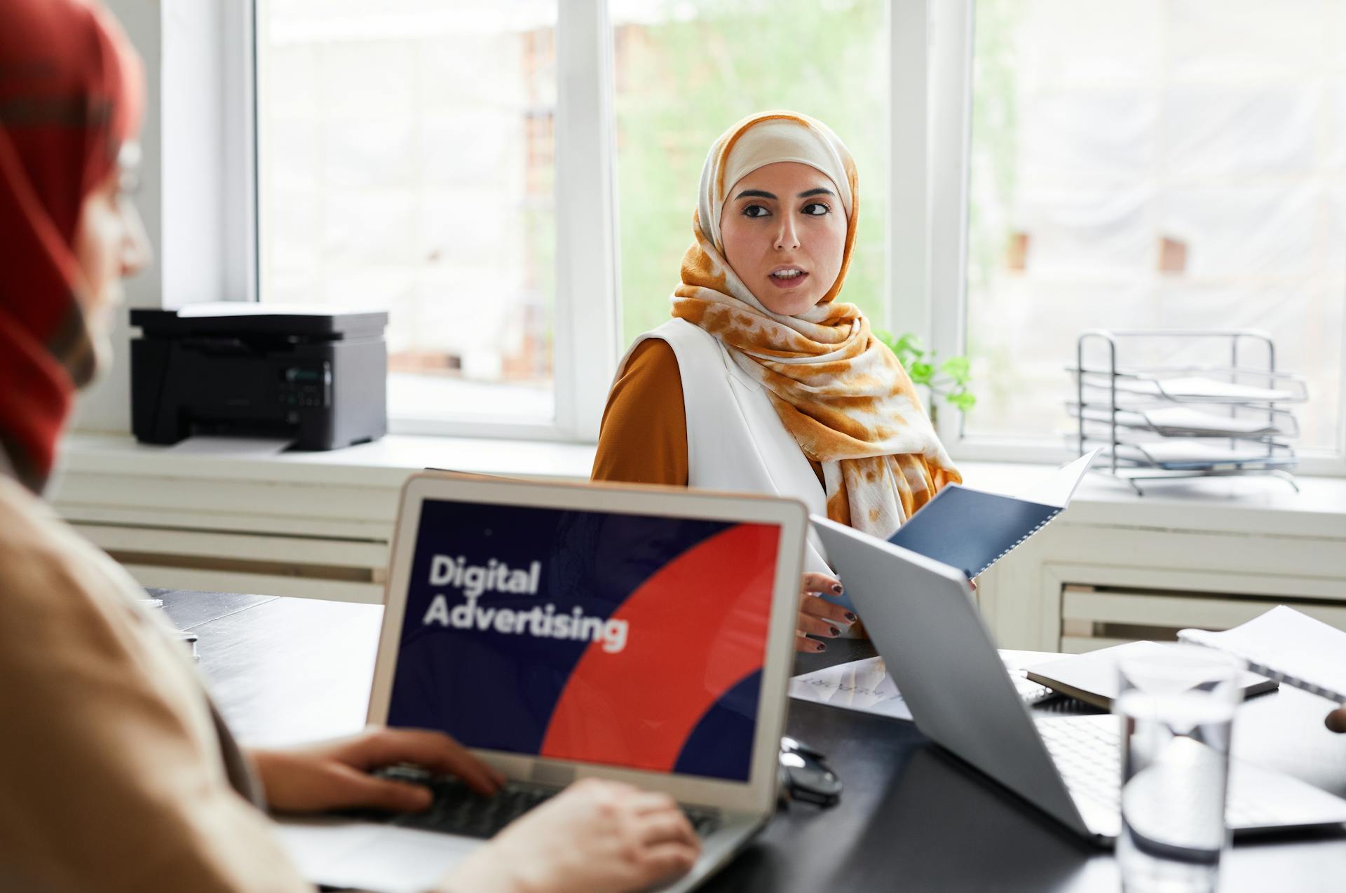 Two women in hijabs discuss digital advertising in an office setting.