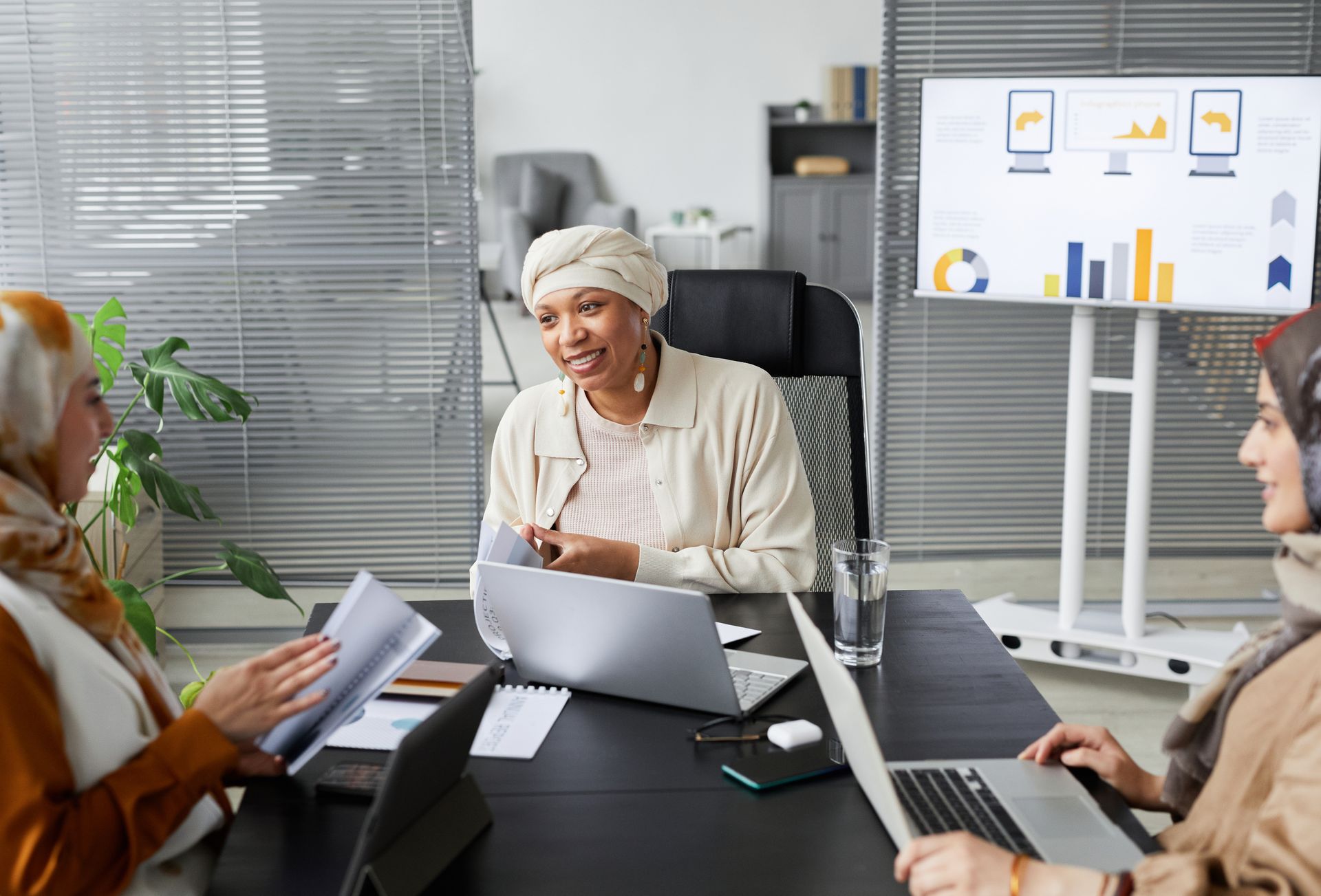 Smiling businesswomen in hijabs at a modern office meeting with laptops and charts.
