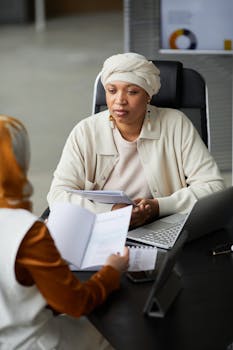 Two women in a business meeting with laptops and documents in a modern office setting.