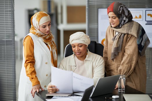 Three professional women in hijabs discussing documents at their office desk. Teamwork and collaboration concept.