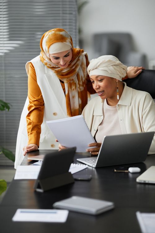 Free Two diverse businesswomen discuss documents together at an office desk. Stock Photo