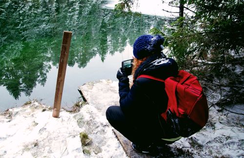 Free Woman with red backpack taking photo by a frozen lake during snowy winter hike. Stock Photo