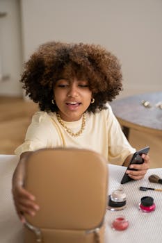 A young woman filming her beauty routine indoors using a smartphone and makeup products.